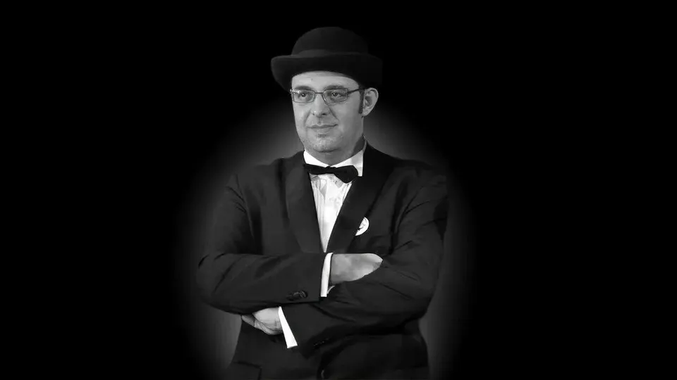 Un bărbat cu ochelari, purtând pălărie, sacou și papion, stă cu brațele încrucișate pe un fundal negru.