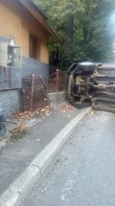 Un autoturism răsturnat stă pe parte pe marginea unui drum, lângă un gard metalic și o clădire cu un panou electric exterior.