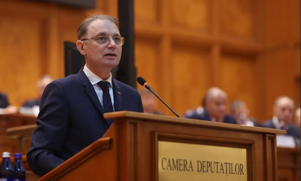 Un bărbat cu ochelari și costum închis la culoare vorbește la un pupitru de lemn pe care scrie Camera Deputaților.