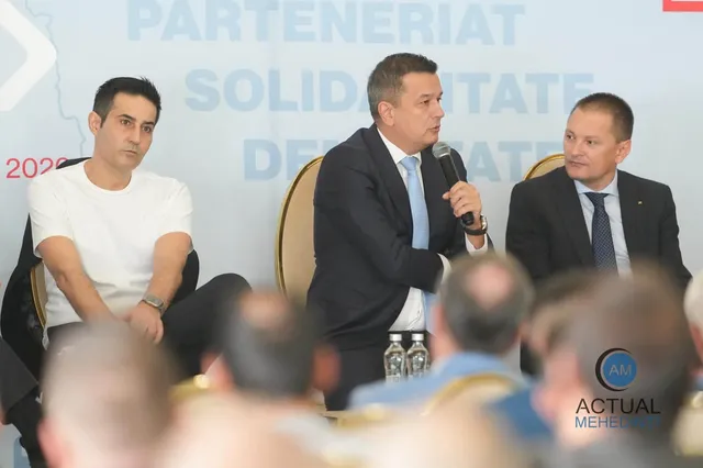 Trei bărbați stau așezați la o masă, unul dintre ei vorbind într-un microfon, în fața unui fundal alb cu text albastru.