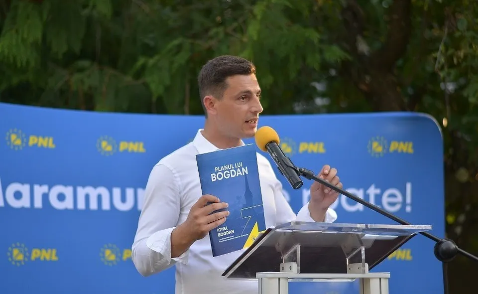 Un bărbat cu cămașă albă ține o carte albastră în mână în fața unui microfon și a unui panou albastru cu text galben.