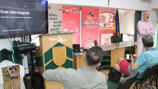 Trei bărbați stau într-o încăpere lângă un pupitru și un ecran de televiziune care afișează text alb pe fundal negru.