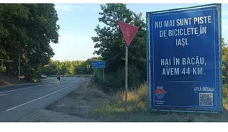 Un panou publicitar albastru cu text alb este amplasat pe marginea unui drum asfaltat lângă vegetație și un semn de circulație triunghiular.