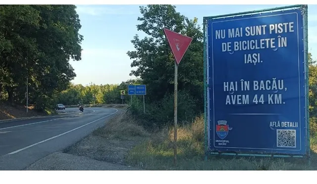 Un panou publicitar albastru cu text alb este amplasat pe marginea unui drum asfaltat lângă vegetație și un semn de circulație triunghiular.
