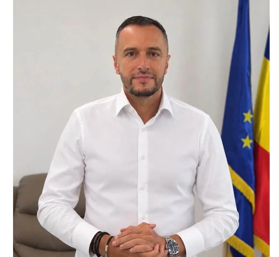 Un bărbat cu barbă scurtă și cămașă albă stă în picioare cu mâinile încrucișate în fața unui fundal alb și a unor steaguri.