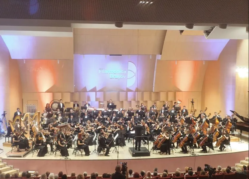 O orchestră de muzică clasică cântă pe o scenă iluminată în fața unui public așezat în sală.
