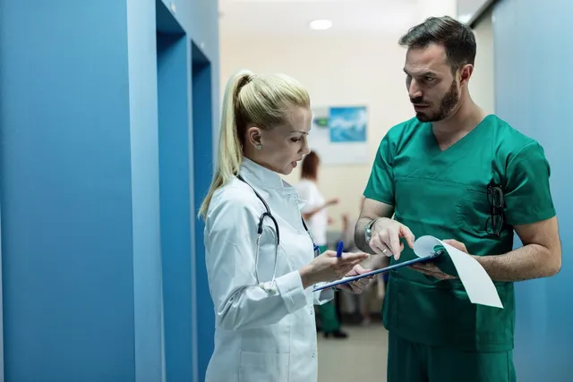 O femeie în halat alb cu stetoscop și un bărbat în echipament verde de medic consultă împreună un dosar pe o clipboard.