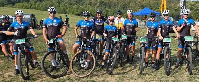 Oameni cu biciclete de munte și căști de protecție stau în rând pe un teren de iarbă sub un cer senin.