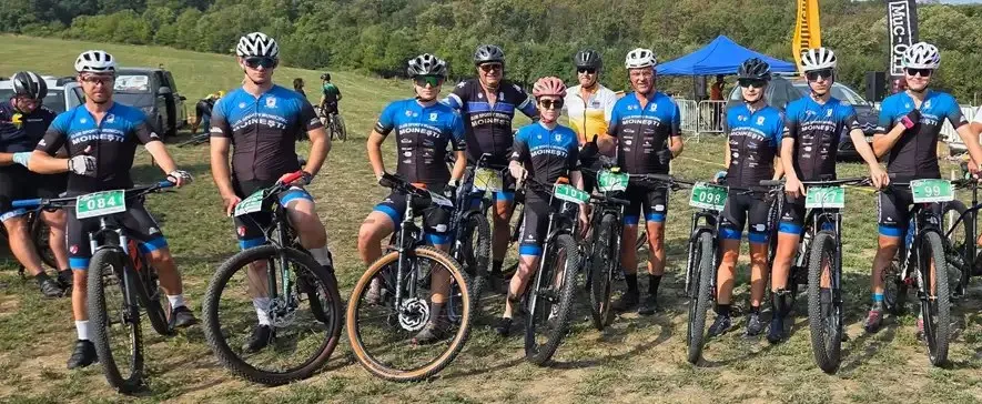 Oameni cu biciclete de munte și căști de protecție stau în rând pe un teren de iarbă sub un cer senin.