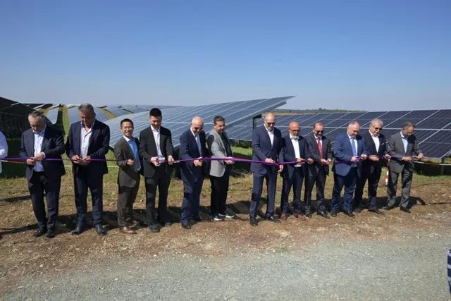 Un grup de bărbați în costume taie o panglică violetă în fața unui câmp cu panouri solare, sub un cer senin.