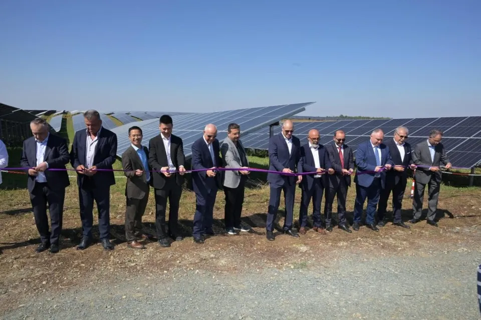 Un grup de bărbați în costume taie o panglică violetă în fața unui câmp cu panouri solare, sub un cer senin.