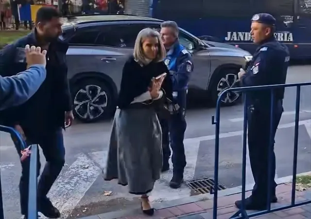 O femeie cu părul blond, purtând o jachetă neagră și o fustă lungă gri, merge pe trotuar lângă un vehicul gri și un polițist.