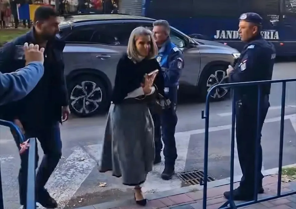 O femeie cu părul blond, purtând o jachetă neagră și o fustă lungă gri, merge pe trotuar lângă un vehicul gri și un polițist.