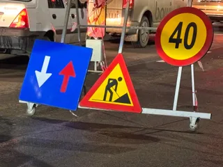Trei semne de circulație pe un suport metal cu roți sunt amplasate pe asfalt lângă două autoturisme albe.