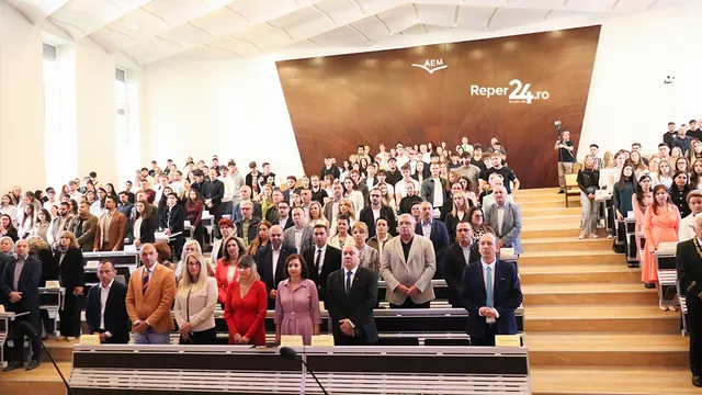 Un grup mare de persoane stă așezat pe scaune în interiorul unei săli de conferințe cu un decor maro în fundal.