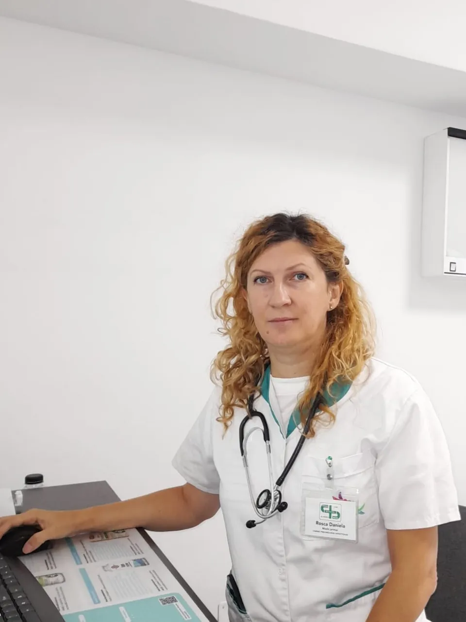 O femeie cu părul creț și bluză albă de medic stă lângă un monitor, purtând un stetoscop în jurul gâtului.