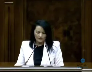 O femeie cu părul negru, purtând o jachetă albă peste un tricou negru, stă în fața unui microfon într-un decor cu panouri de lemn.