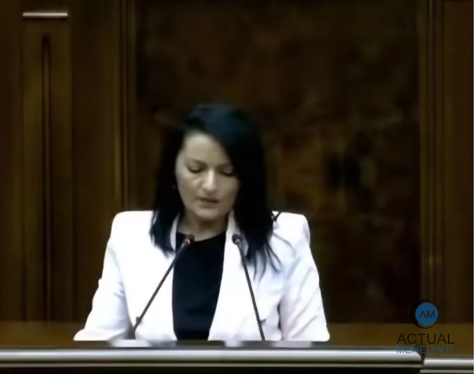 O femeie cu părul negru, purtând o jachetă albă peste un tricou negru, stă în fața unui microfon într-un decor cu panouri de lemn.