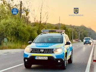 Un autovehicul de poliție albastru și alb circulă pe un drum asfaltat cu vegetație și un alt vehicul în fundal.