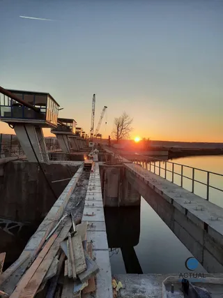 O structură de beton cu parapet metalic se întinde peste apă, având macarale și un apus de soare în fundal.
