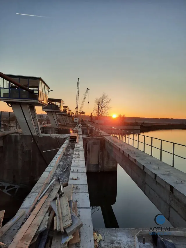 O structură de beton cu parapet metalic se întinde peste apă, având macarale și un apus de soare în fundal.