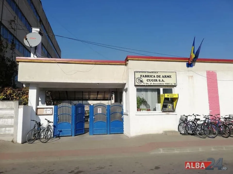 O clădire albă cu un gard albastru, un afiș cu text și câteva biciclete parcate în fața acesteia sub un cer senin.