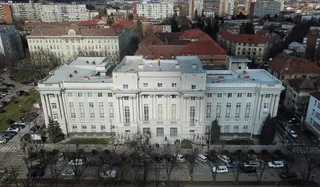 O vedere aeriană a unei clădiri albe cu ferestre multiple, înconjurată de alte construcții urbane și copaci fără frunze.