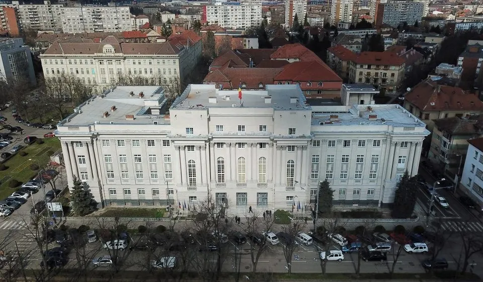 O vedere aeriană a unei clădiri albe cu ferestre multiple, înconjurată de alte construcții urbane și copaci fără frunze.