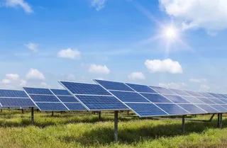O instalație de panouri solare fotovoltaice este amplasată pe un teren cu iarbă verde, sub un cer albastru însorit.