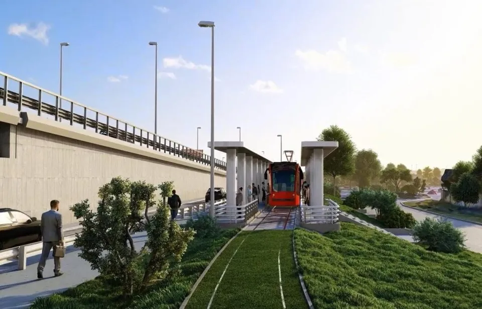 Un tram-tren roșu și negru circulă pe o linie de rulare lângă o stație cu acoperiș și persoane pe un trotuar.