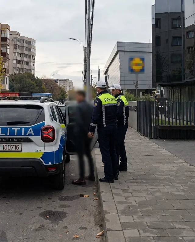 Unii polițiști în uniformă stau lângă o mașină albastră și albă marcată cu textul POLIȚIA pe un trotuar lângă un clădire.