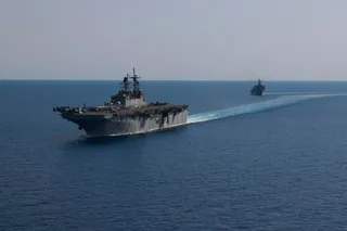 Două nave militare gri navighează pe o suprafață de apă albastră sub un cer senin.
