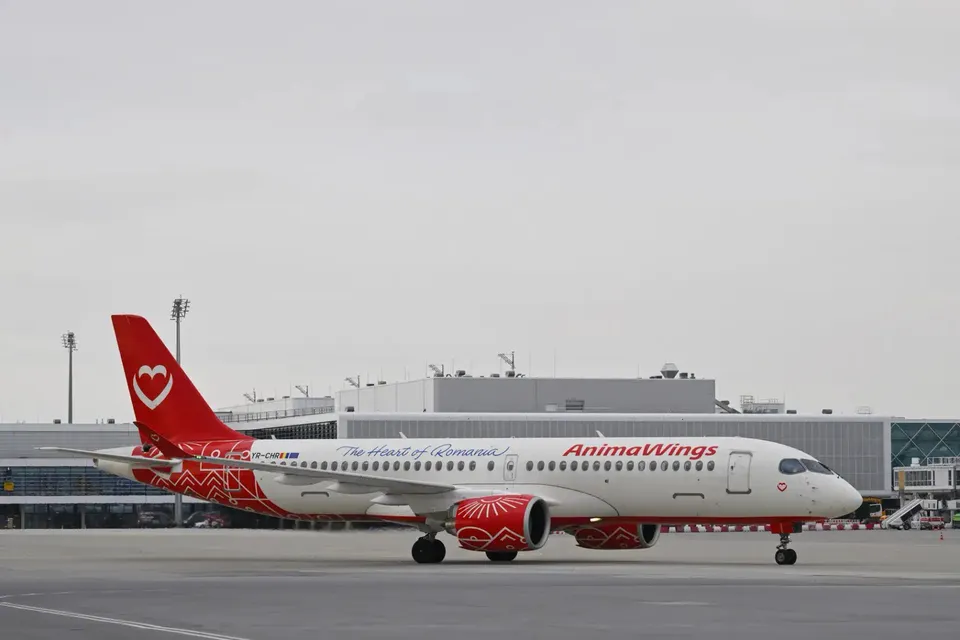 Un avion alb cu accente roșii și inscripția AnimaWings stă pe o pistă de aeroport sub un cer gri.