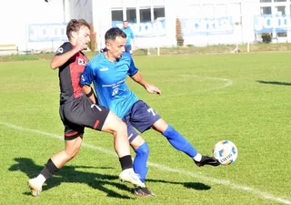 Un jucător în echipament albastru întinde piciorul spre un fotbal pe un teren de fotbal verde, fiind urmărit de un jucător în echipament negru și roșu.