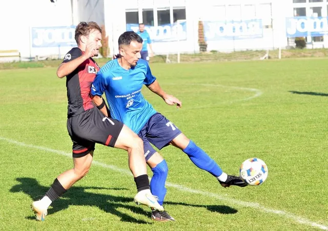 Un jucător în echipament albastru întinde piciorul spre un fotbal pe un teren de fotbal verde, fiind urmărit de un jucător în echipament negru și roșu.