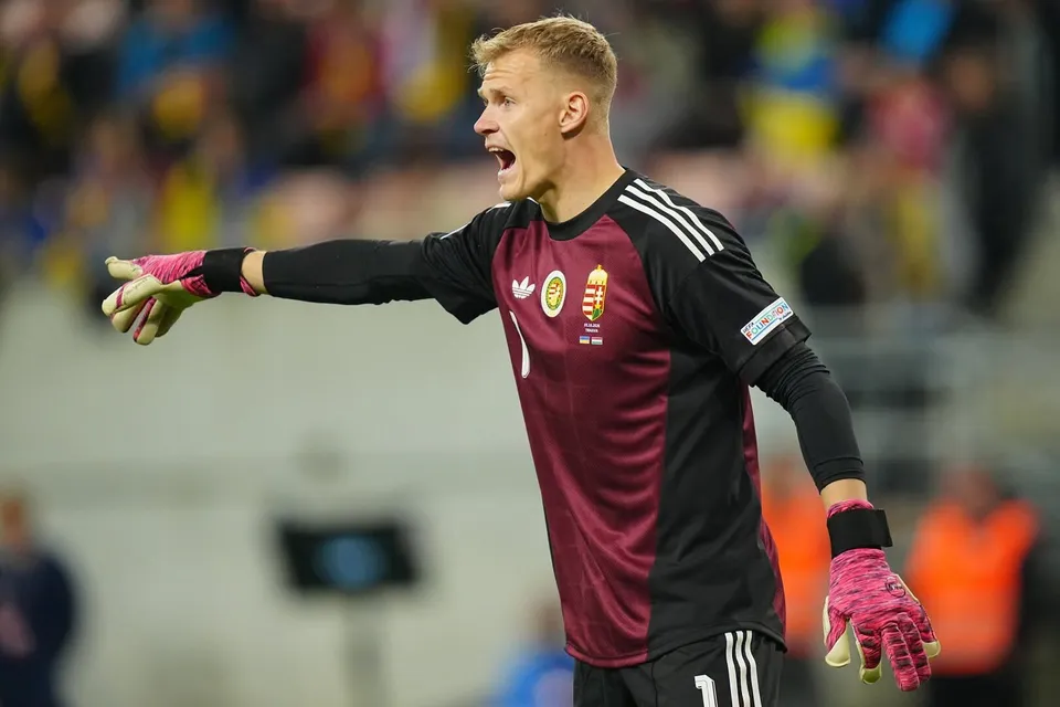 Un fotbalist cu mănuși de portar, purtând un tricou negru și bordo, stă în picioare și arată cu mâna stângă spre lateral.