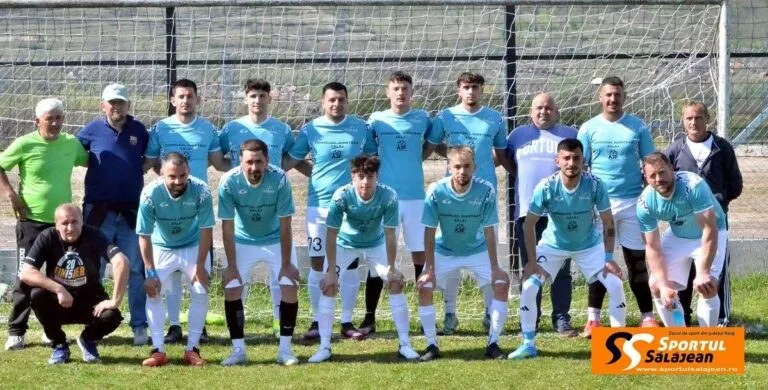 O echipă de fotbaliști în echipamente albastre și albe pozează în fața unei porți de fotbal pe un teren de iarbă.