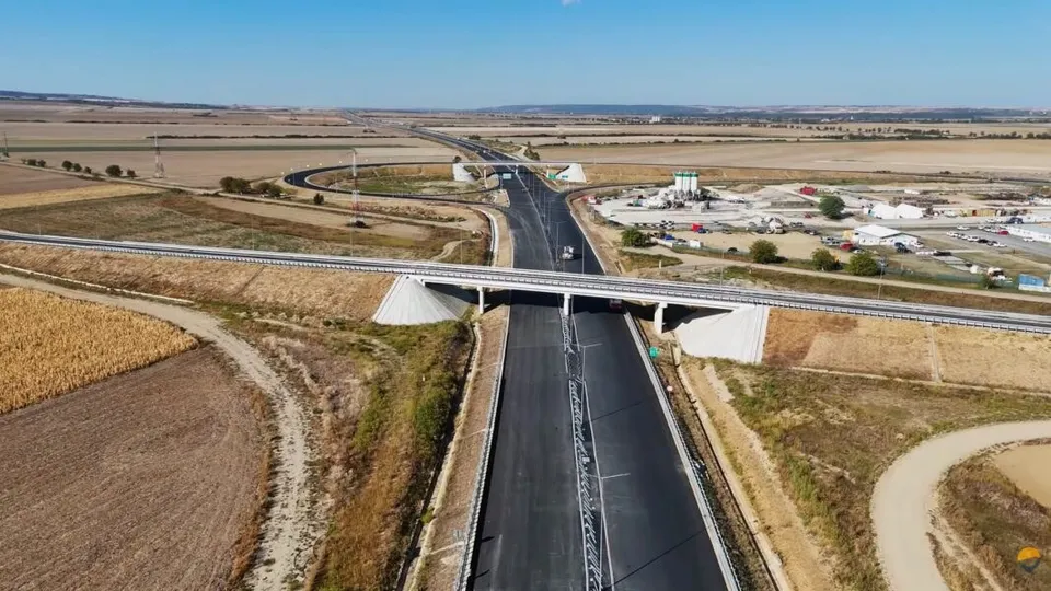 O autostradă cu două benzi de fiecare sens traversează un peisaj agricol plat, fiind traversată de un pod de o altă cale de rulare.