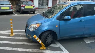 Un autovehicul albastru este parcat peste marcajele de trecere de pietoni, lovind un stâlpișor galben și alb.