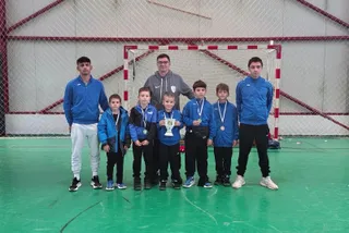Un grup de băieți în vârsta de școală, însoțiți de doi adulți, pozând cu medalii la gât pe un teren de sport interior.