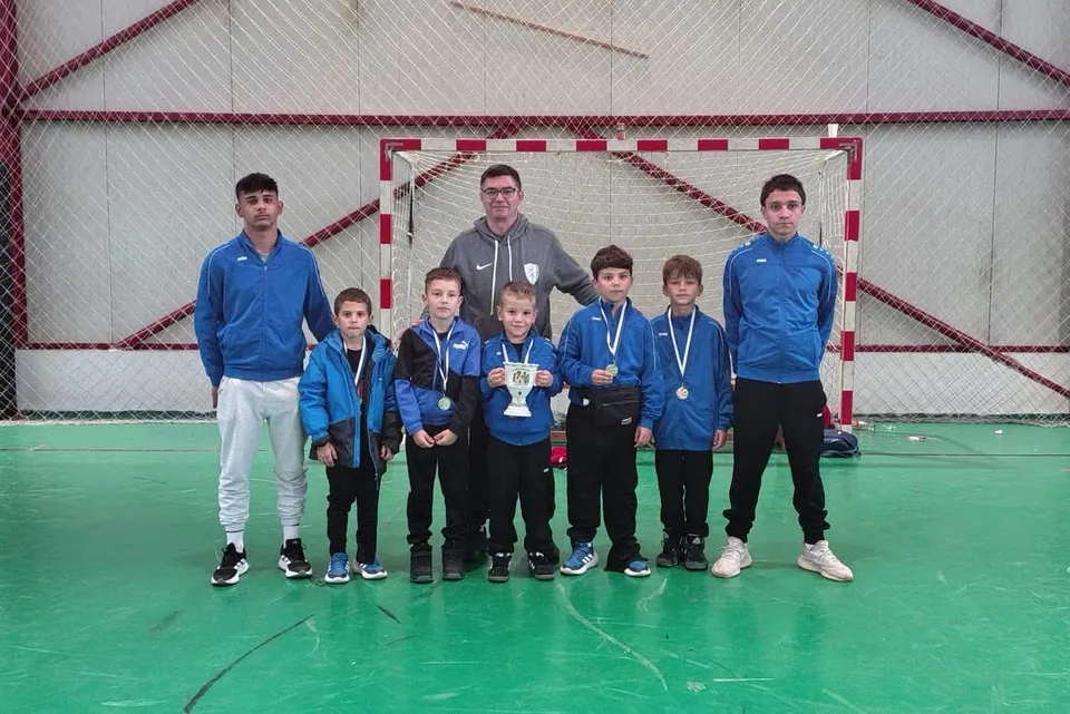Un grup de băieți în vârsta de școală, însoțiți de doi adulți, pozând cu medalii la gât pe un teren de sport interior.