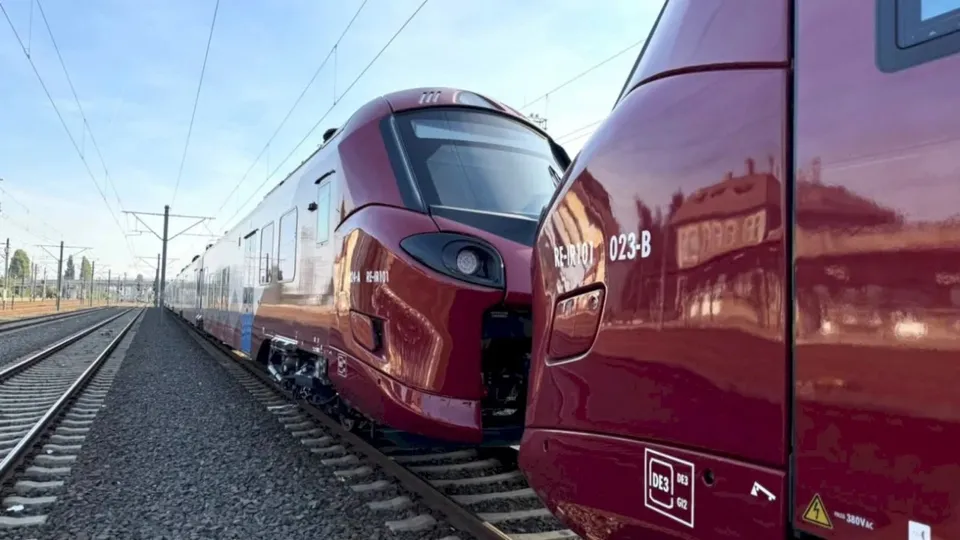 Două rame electrice de culoare vișinie sunt amplasate pe șine de cale ferată sub un cer senin.