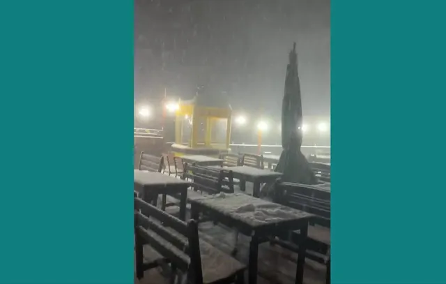 Mese și bănci de lemn acoperite de zăpadă sunt situate pe o terasă exterioară sub o precipitație de zăpadă în timpul nopții.