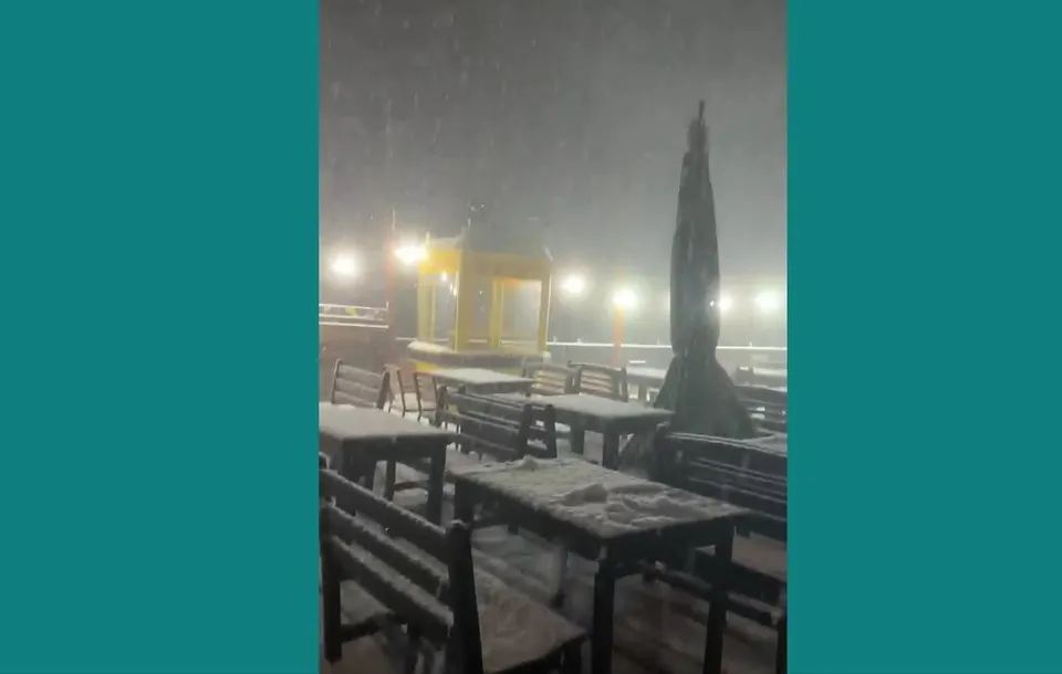 Mese și bănci de lemn acoperite de zăpadă sunt situate pe o terasă exterioară sub o precipitație de zăpadă în timpul nopții.