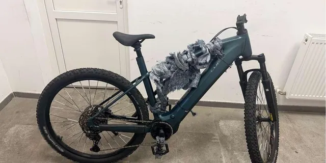 O bicicletă de munte de culoare închisă este oprită în profil într-o încăpere cu un calorifer și o ușă albă în fundal.