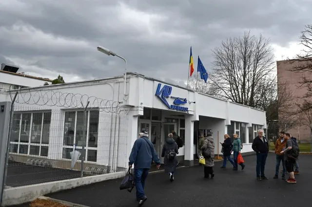 Oamenii merg pe o stradă asfaltată în fața unei clădiri albe cu ferestre mari, sub un cer înnorat.