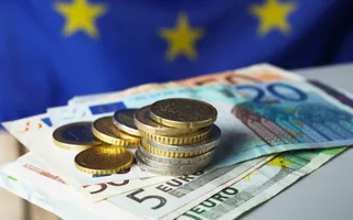 Monede metalice sunt așezate pe bancnote de euro, în fața unui steag albastru cu stele galbene.