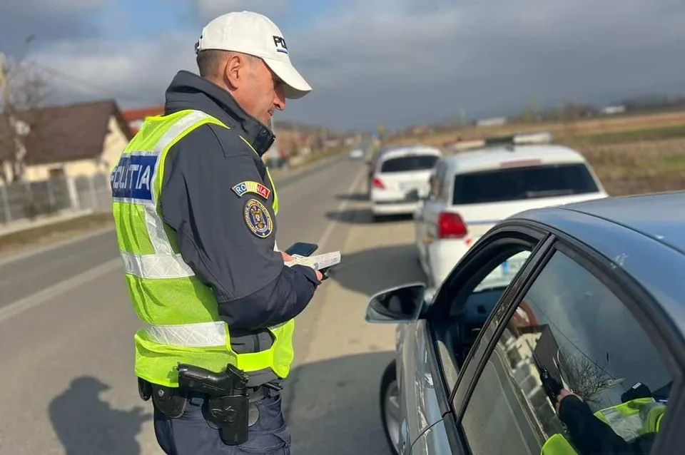 Un polițist în uniformă și vestă reflectorizantă verifică documentele unui șofer aflat într-o mașină de culoare gri pe marginea unui drum.