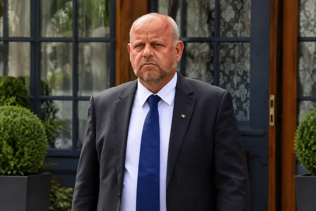 Un bărbat cu părul scurt și barbă stufoasă poartă un costum închis, cămașă albă și cravată albastră în fața unei ferestre cu rame negre.