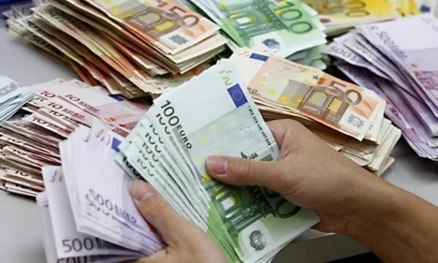 Mâinile unui individ țin un teanc de bancnote de o sută de euro deasupra unor alte teancuri de bancnote de diverse valori.
